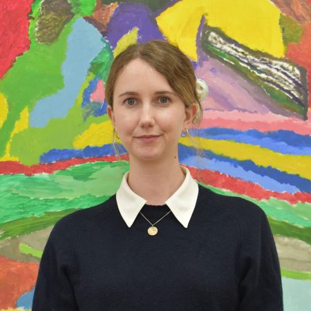 A picture of a white woman with brown hair that's been tied behind her head. She is wearing a dark top with a white collar and is also wearing a necklace. She is standing in front of a painting.