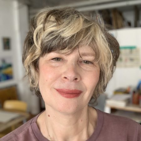 A white woman with short blonde hair, red lipstick and is wearing a brown t shirt.