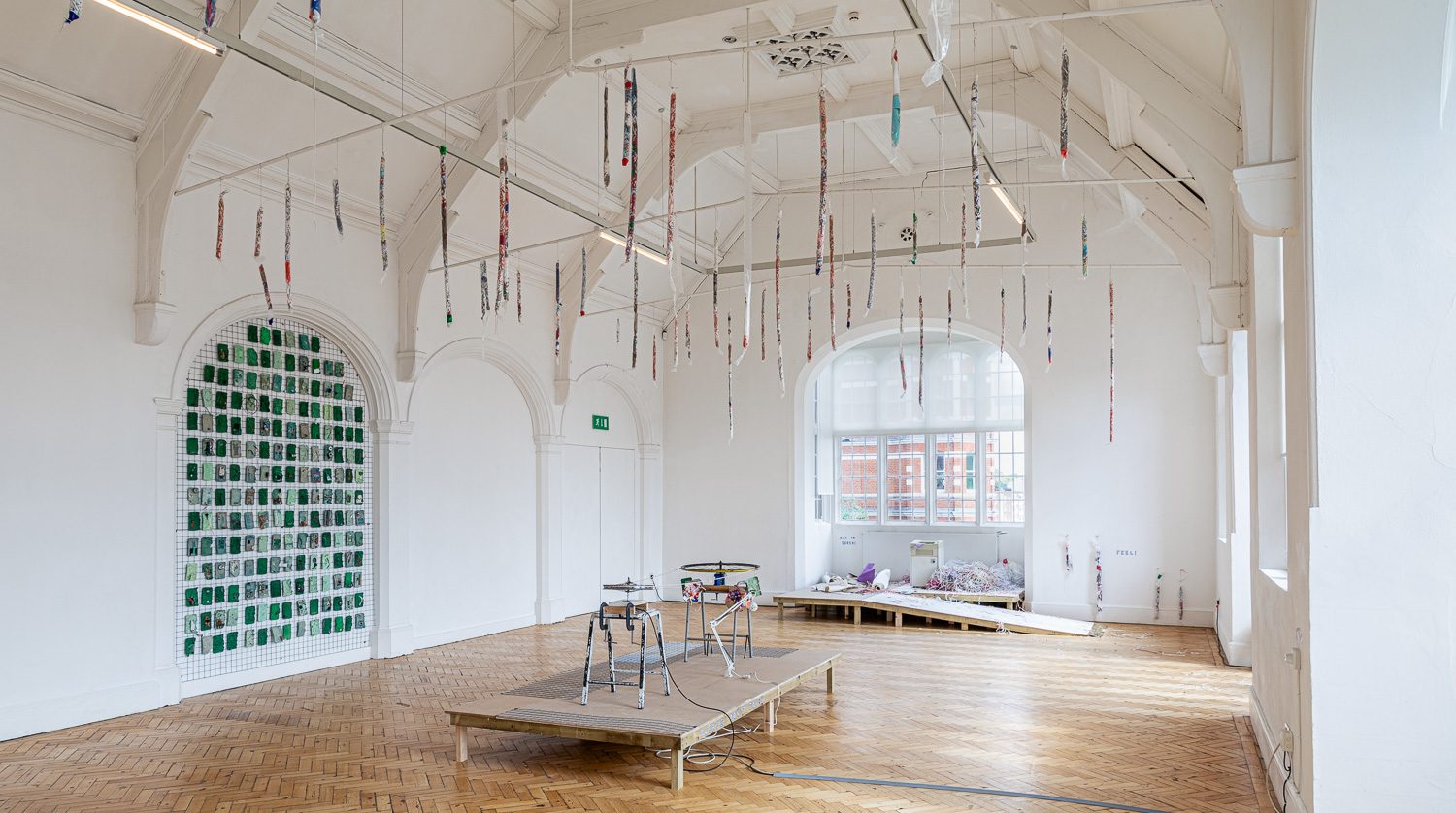 A gallery space with thin fabric strips suspended from three railings attached to the ceiling. In an arch, small green artworks fill in its cavity. The floor of the gallery has two short platforms with assemblage sculptures on top.