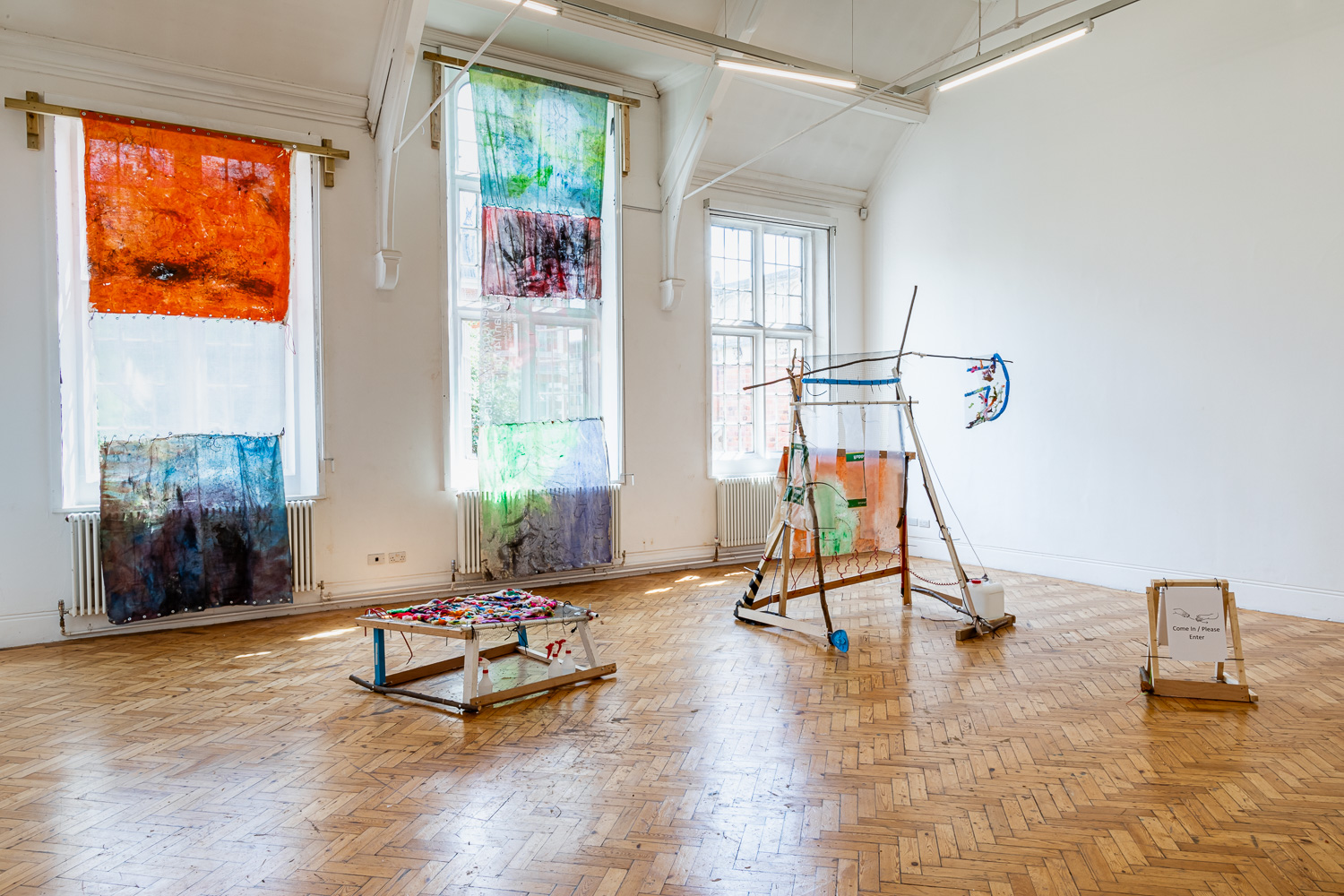 An exhibition space with two hand dyed fabric pieces hanging in front of the windows and three wooden sculptures.