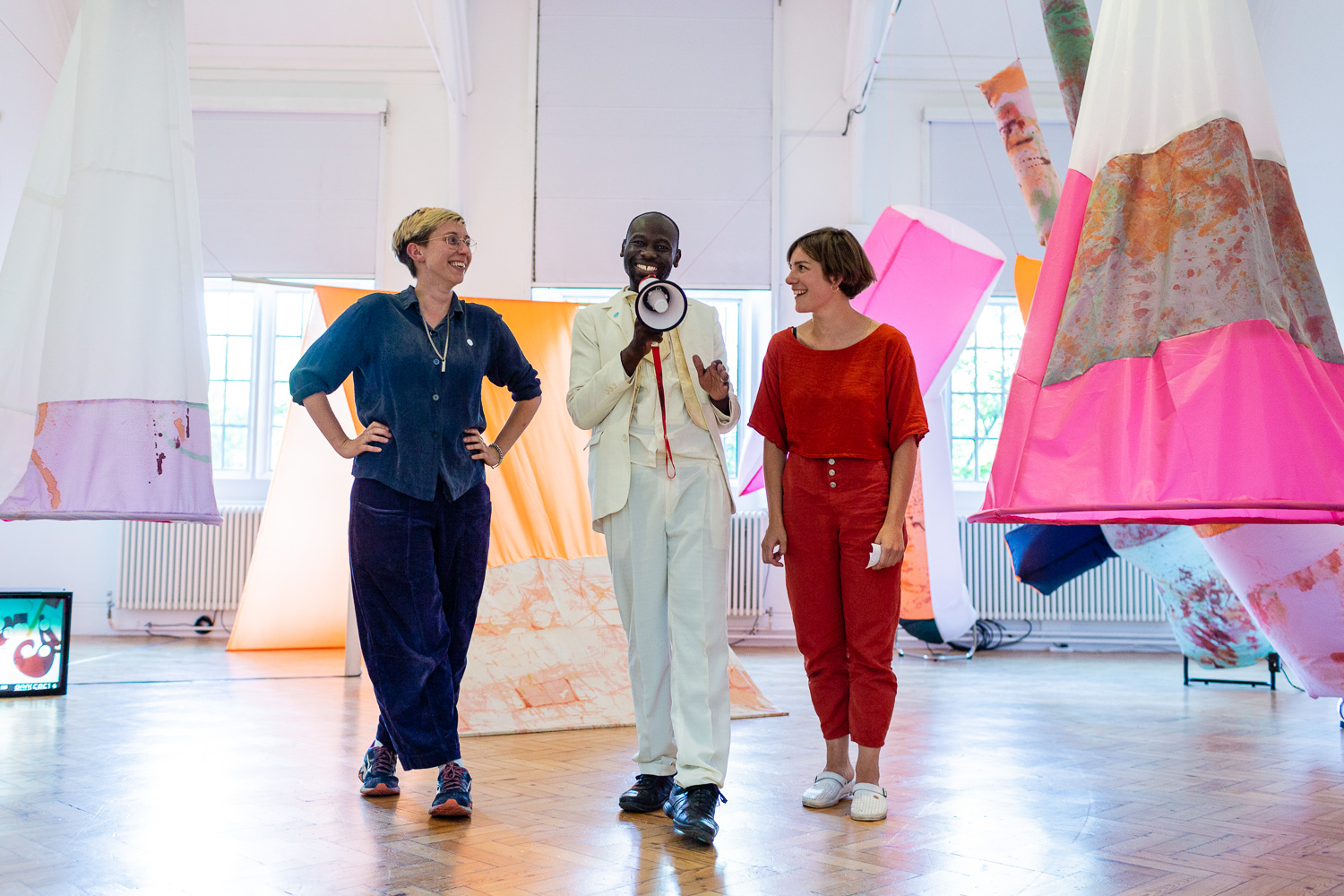 Artist Andrew Omoding, speaking into a megaphone, stood between two women in front of an exhibition with six tent structures hung up in a gallery space. A CRT TV, playing a video, sits on the floor.