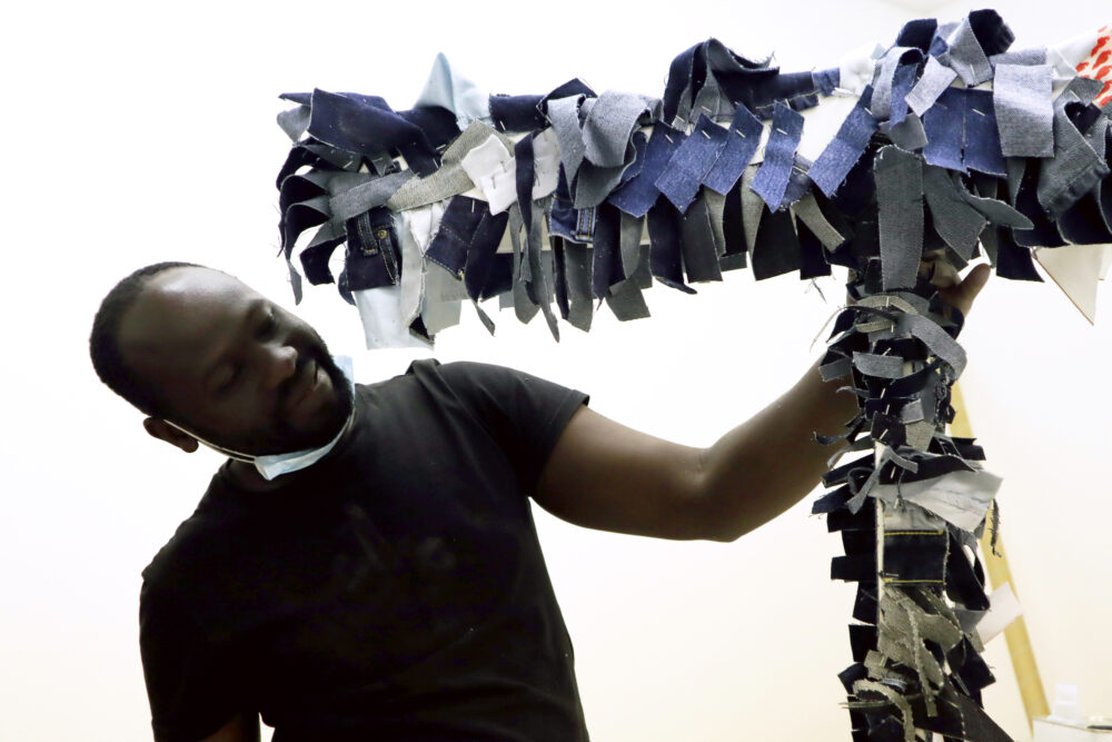 Andrew holds one of his sculptural artworks. It is a post with lots of small fabric denim strips stapled to it.