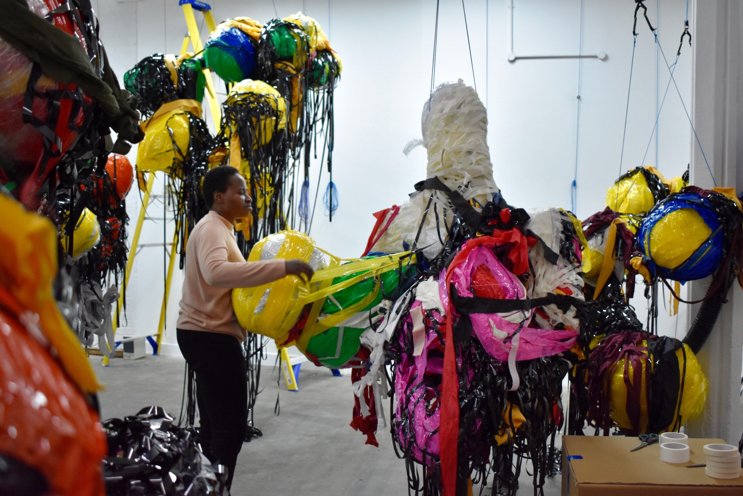 Nnena Kalu standing and building a boulder onto her suspended artworks make out of layers of tape, paper and plastic.