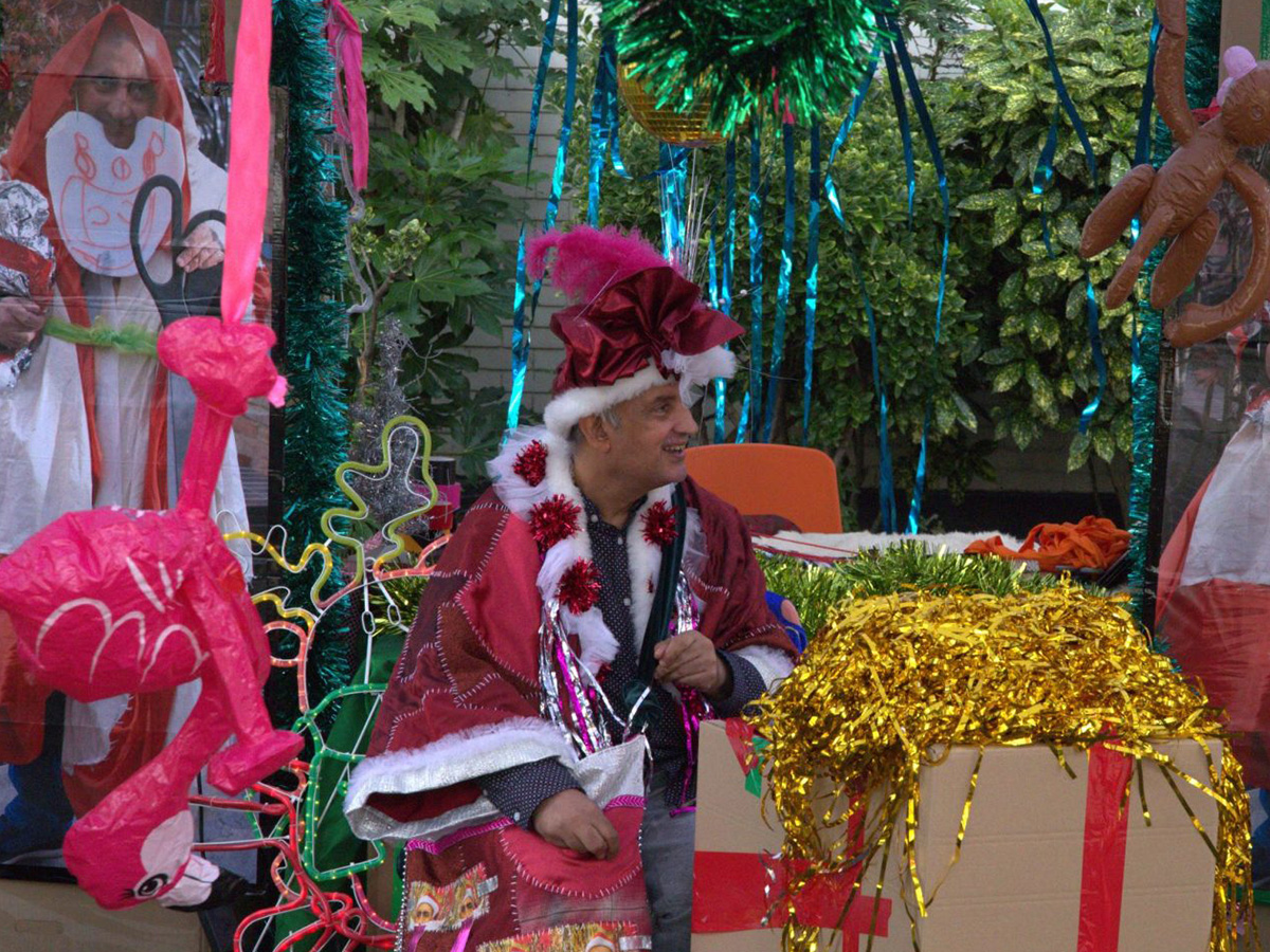 Pardip Kapil sits surrounded by his artwork. He is wearing a handmade santa outfit, and sits near to gold tinsel and an inflatable pink flamingo.