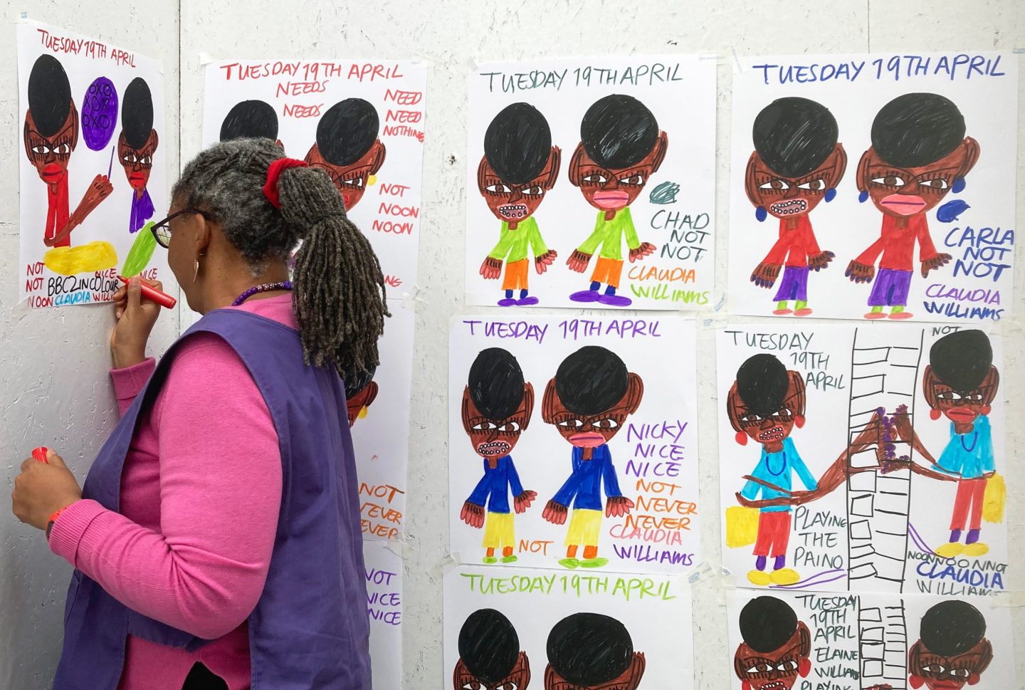 Claudia Williams stands near a wall, drawing on one of her artworks which is fixed to the wall. A selection of her other artworks are also on the wall, showing felt tip drawings of vinyl covers.
