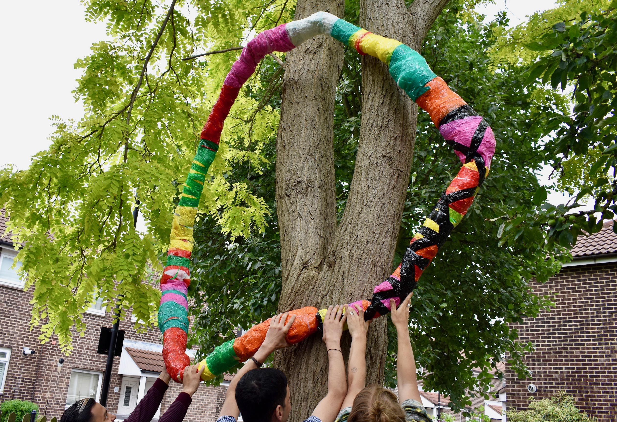 3 people holding up a colourful hoop sculpture against a tree.