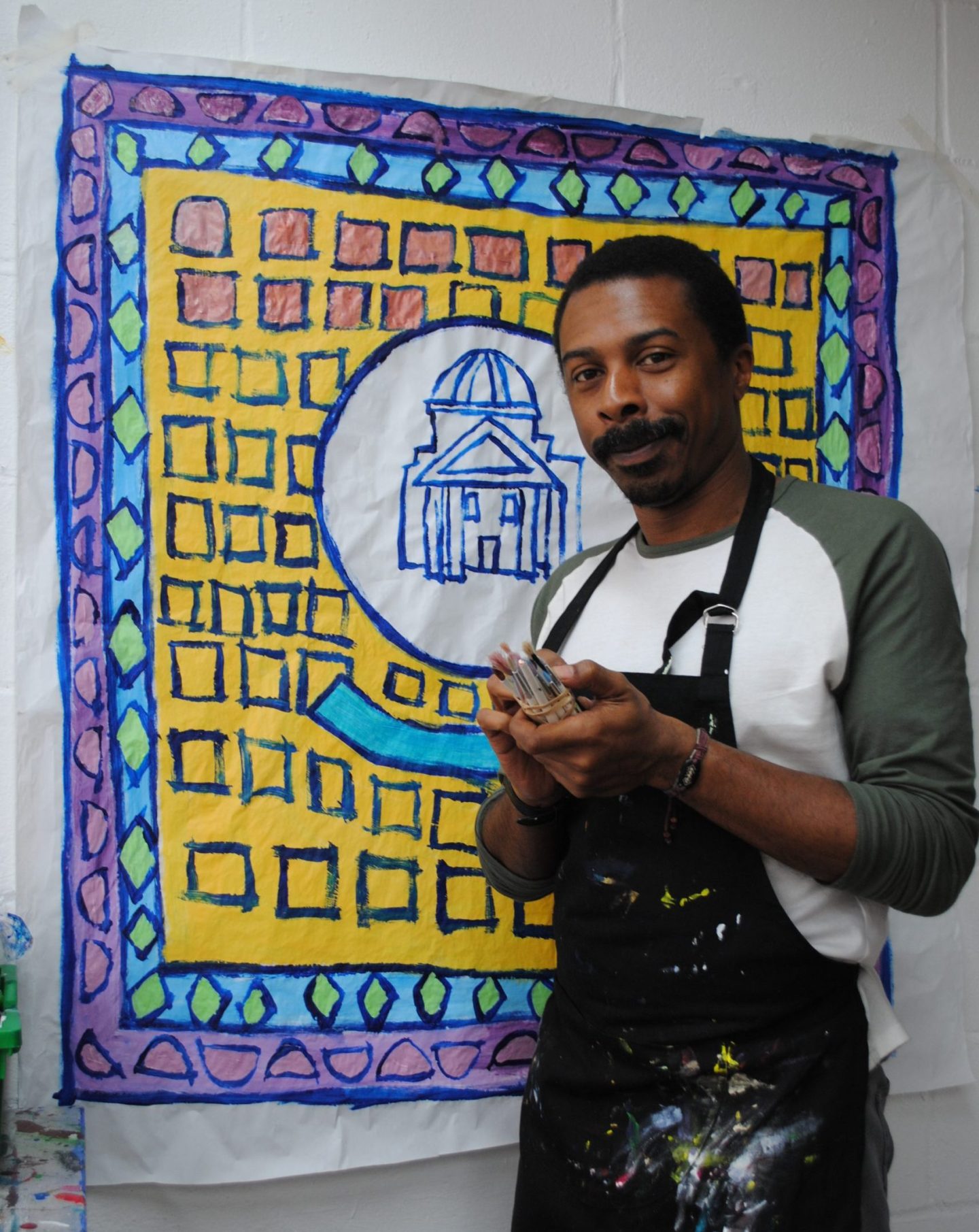 Thompson Hall stands in front of his artwork holding paintings in his hands.