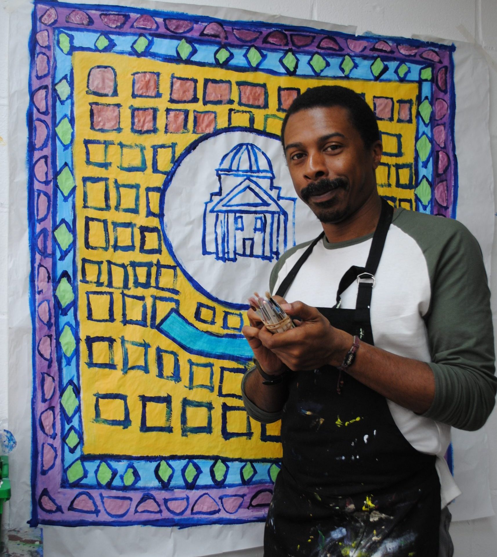 Thompson Hall stands in front of his artwork holding paintings in his hands.