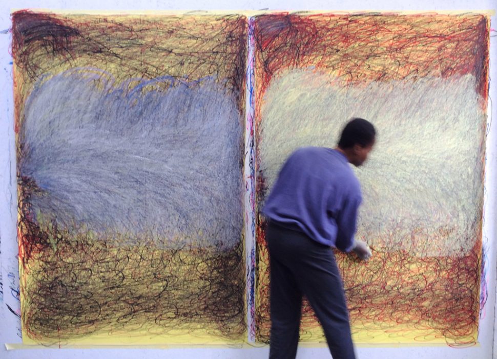 Nnena Kalu works in front of two large pieces of hers. There are two large yellow pieces of paper, with scrawled dark black and red marks on them, and a large strip of blue and white in the middle.