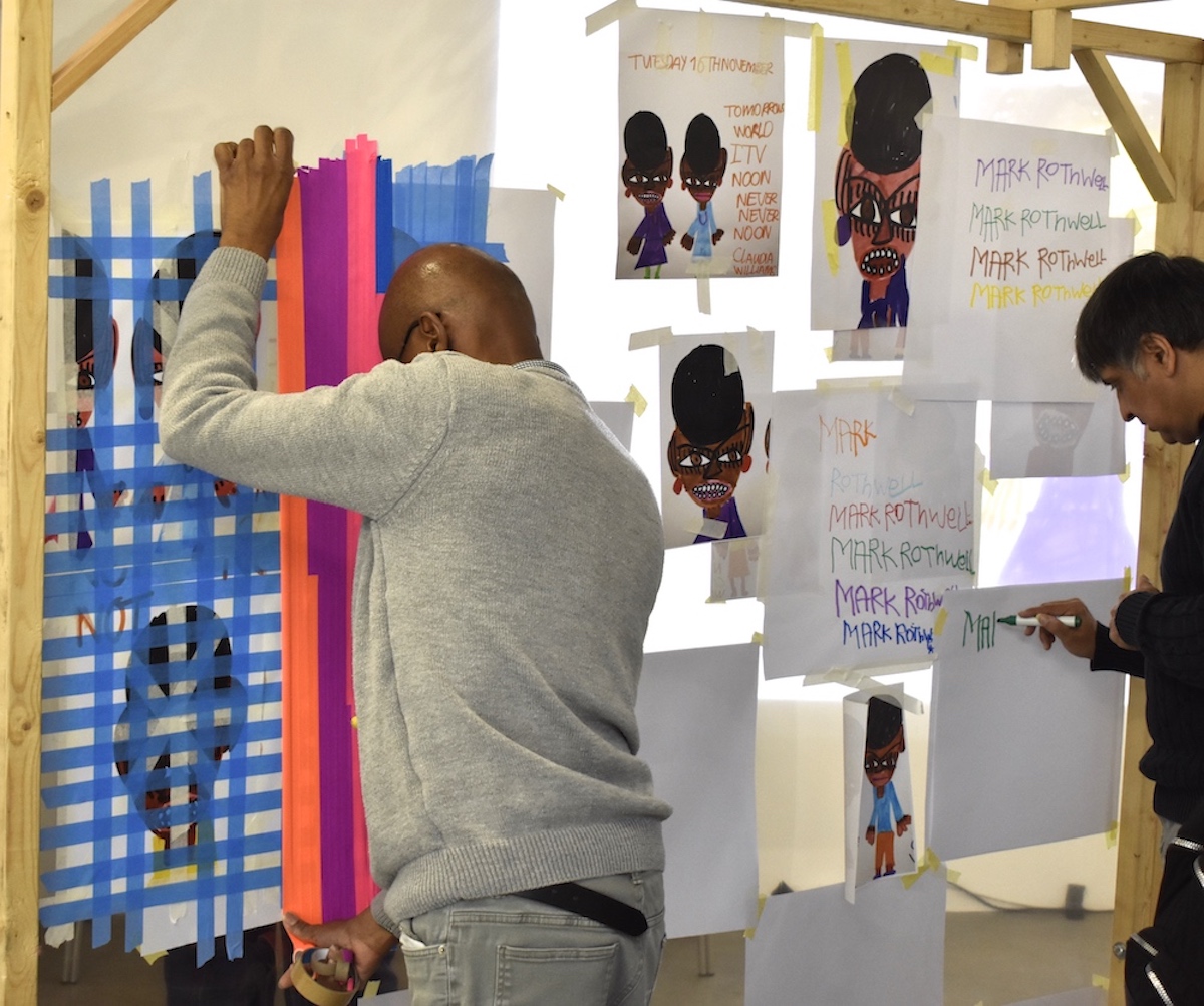 On a large wooden frame, one man is taping over artwork with layers of colourful tape. Another man is writing a name repeatedly on paper.