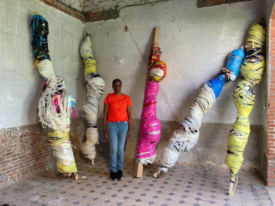 Nnena Kalu stands against a brick wall, with 5 of her very large sculptures resting on the wall surrounding her. She is half the size of the sculptures which are very tall, and are colourful wound and taped artworks.