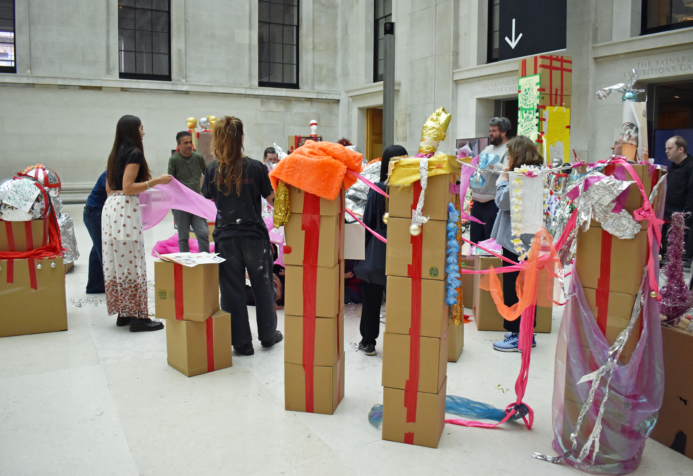 A crowd of participants stand in between sculptures made of stacked cardboard boxes, tape and found textile pieces. They are holding a pink, sheer fabric out like a parachute.