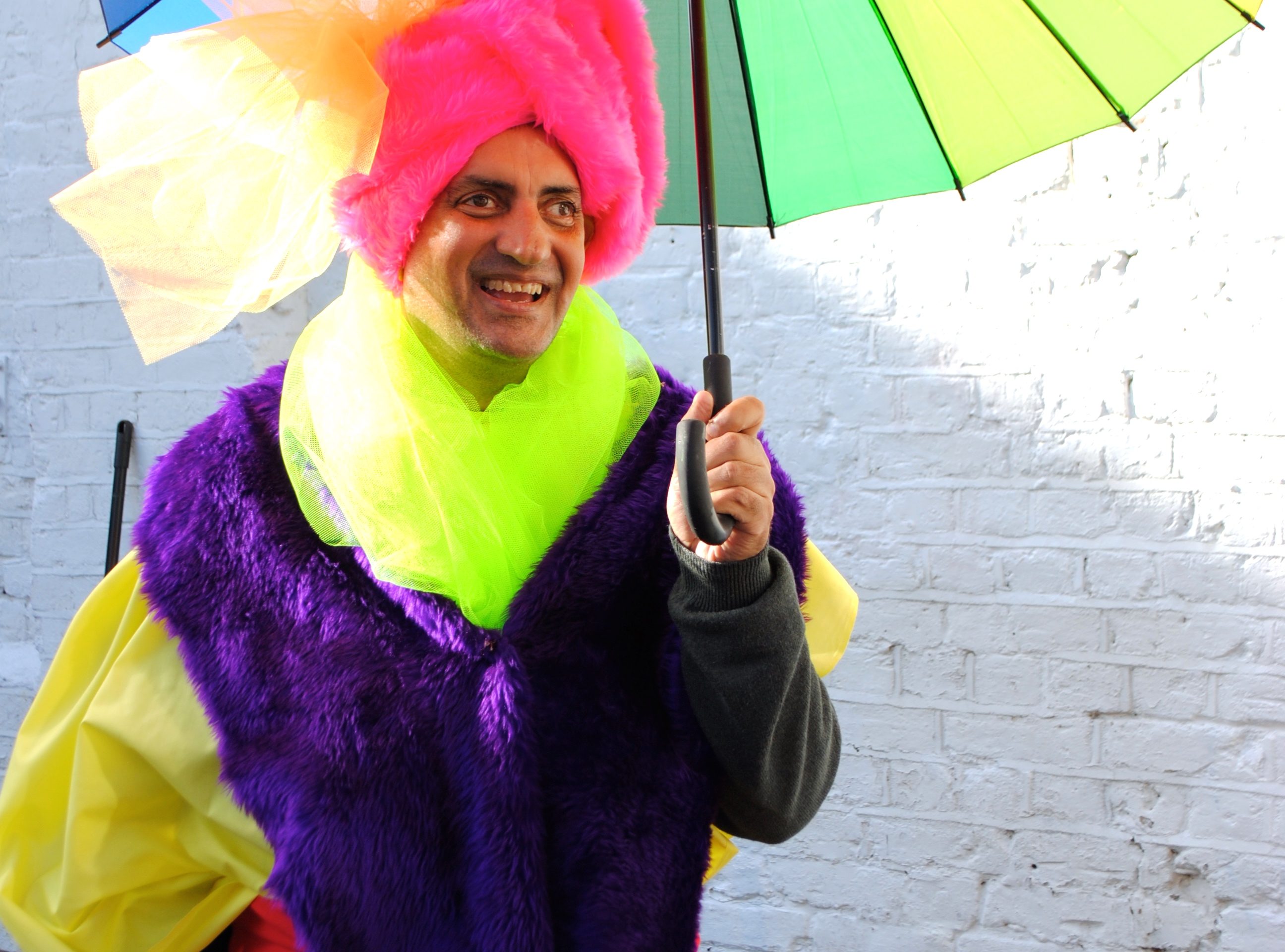 Pardip holds a rainbow umbrella and smiles. He is wearing a bright pink turban, a neon yellow scarf, a furry purple piece of fabric over his shoulders and a yellow jacket underneath.
