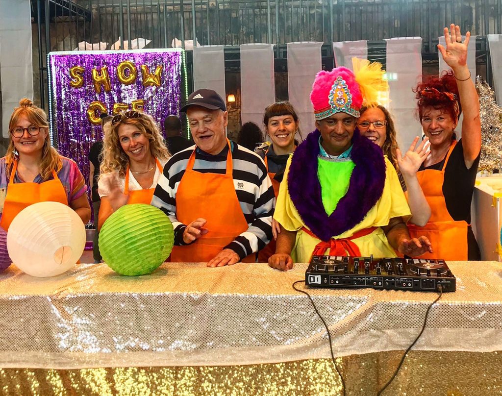 7 people, a mix of ActionSpace artists and staff, stand behind a table. There is a DJ deck on the table and two round light shades. Everyone is smiling and wearing aprons.
