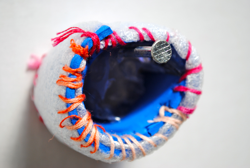 A small textile artwork shaped like a hollow cylinder hanging from a nail on a wall. The white outer fabric is lined with blue, and it is joined together by different coloured overcast stitches.