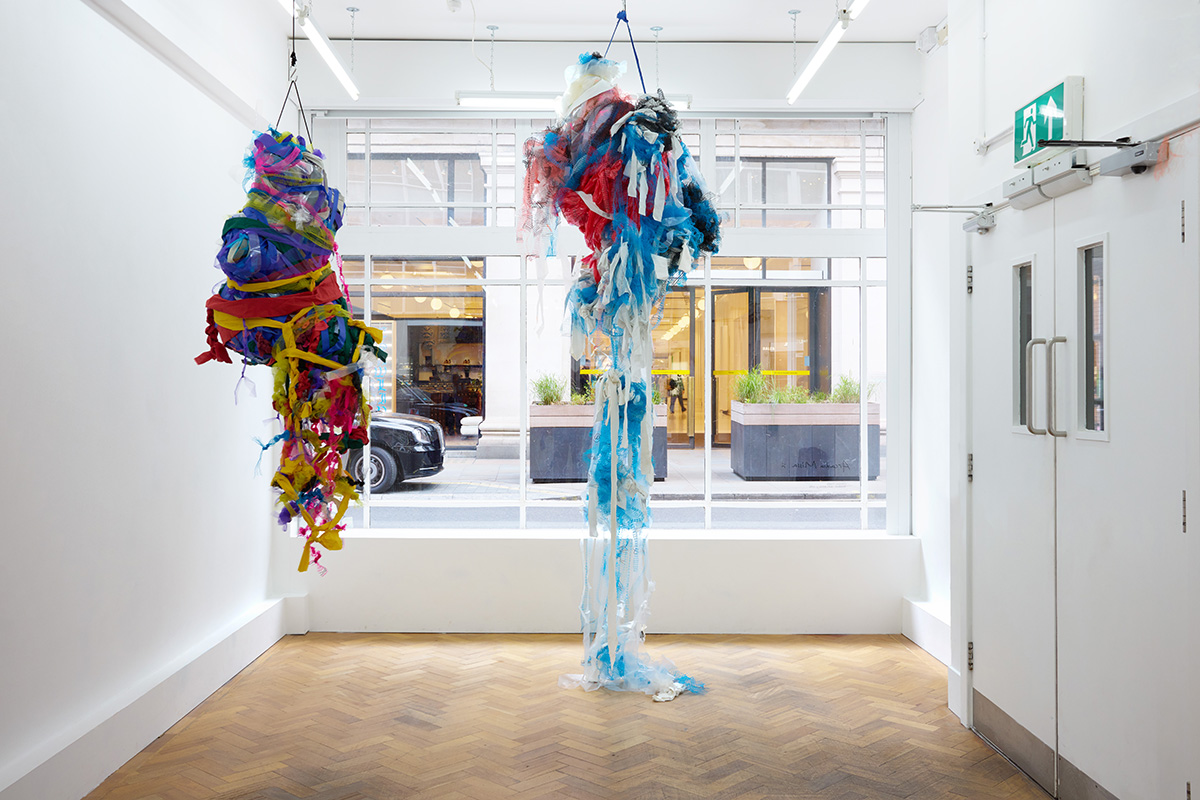 Two colourful, large mixed media sculptures hung from the ceiling in front of a gallery window, with one trailing on the floor.