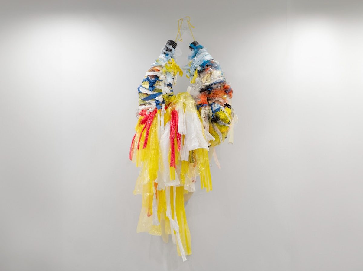 A colourful hanging sculpture made of looped strips of differing materials, with yellow, white and pink tail ends hanging downwards.