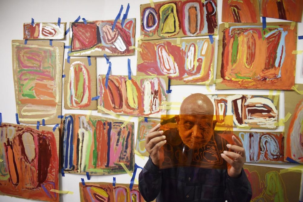 Chandrakant looks through a transparent sheet of orange accetate. Behind him are a selection of his works on the wall.