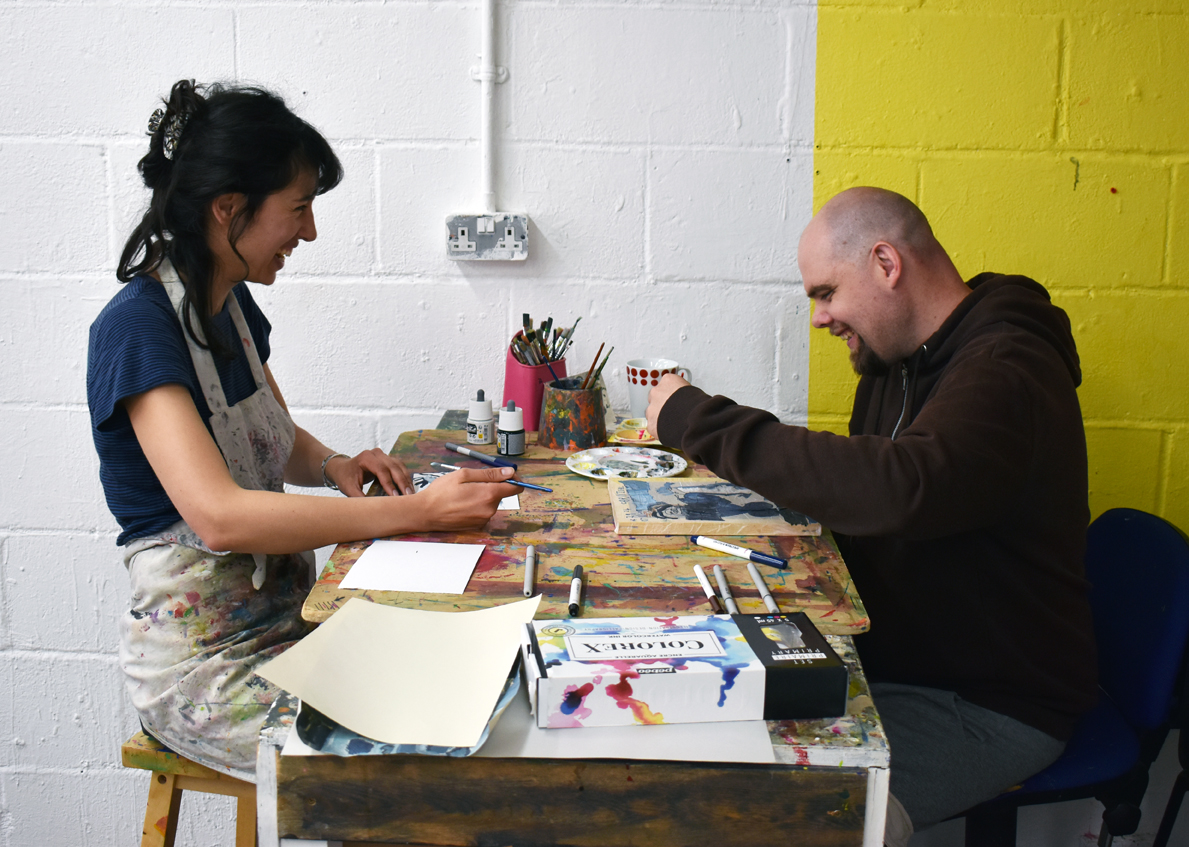 Artist Declan Leslie working in the studio at his table with a volunteer.