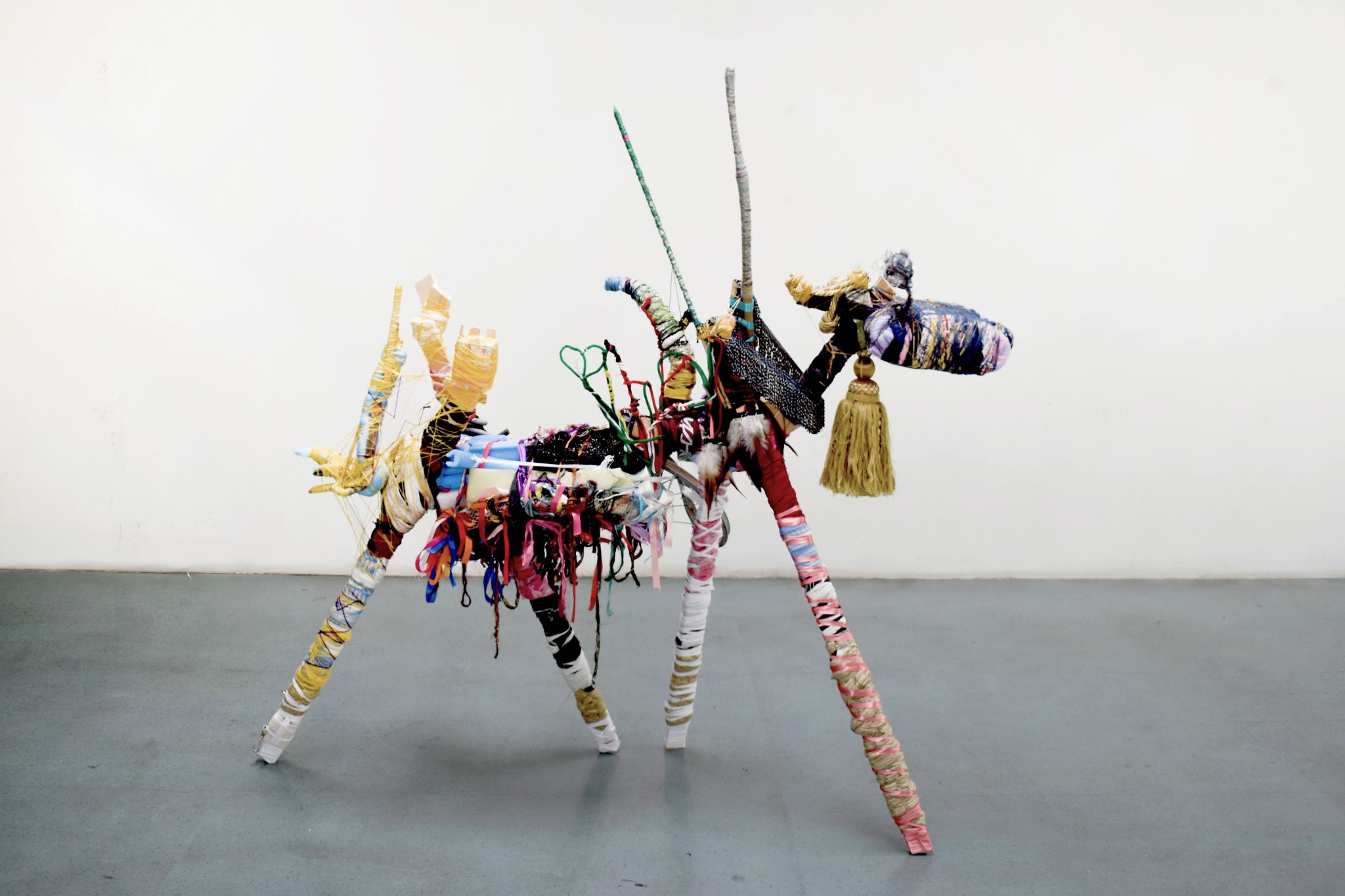 A mixed media sculpture stood on a grey concrete floor in a white walled room. The sculpture, resembling a goat, is made of various materials such as pink ribbon, gold tassel, blue foam etc.