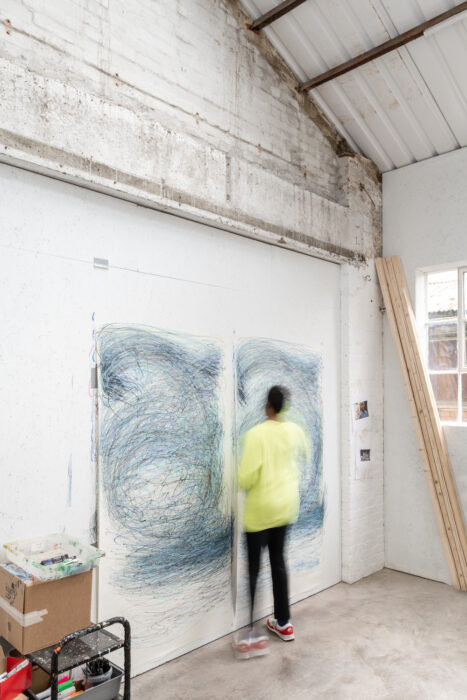 Nnena Kalu working in her studio. She stands drawing on two large pieces of white paper, making blue spirals.