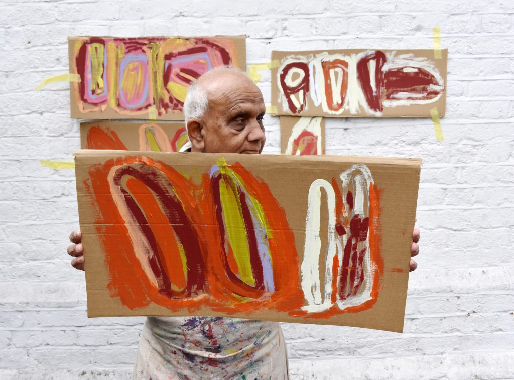 Chandrakant holds his work up to the camera. It is an abstract, painted piece done on cardboard. There is orange, red and white circles in the painting.
