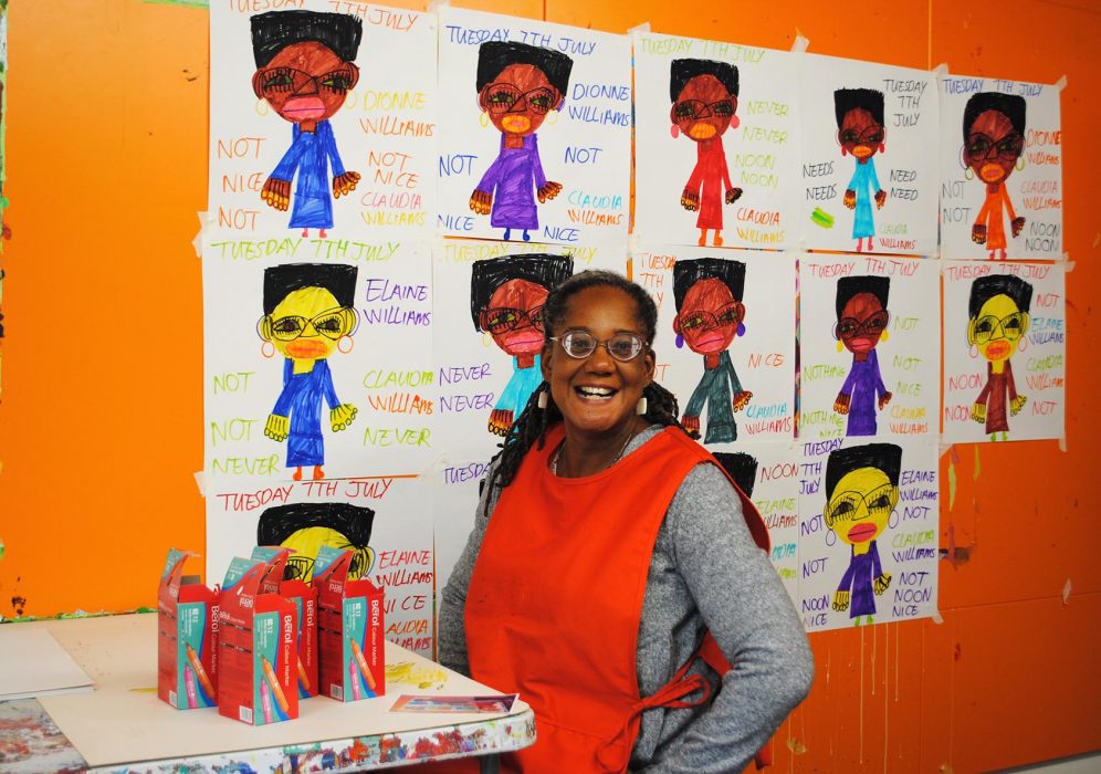 Claudia Williams artist portrait. She is standing in front of an orange wall, wearing an apron and smiling at the camera. Behind her is her work that is displayed on the wall.