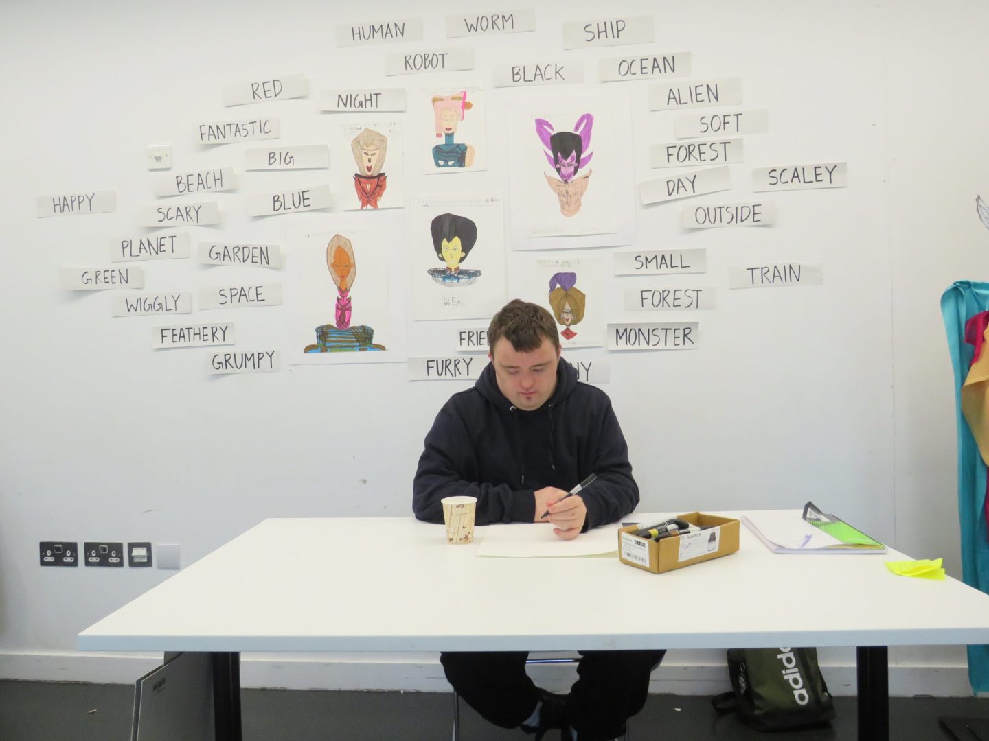 Artist Benjamin Allen sits at a table ready to draw during one of his workshops. His character illustrations and text prompts are on the wall behind him.