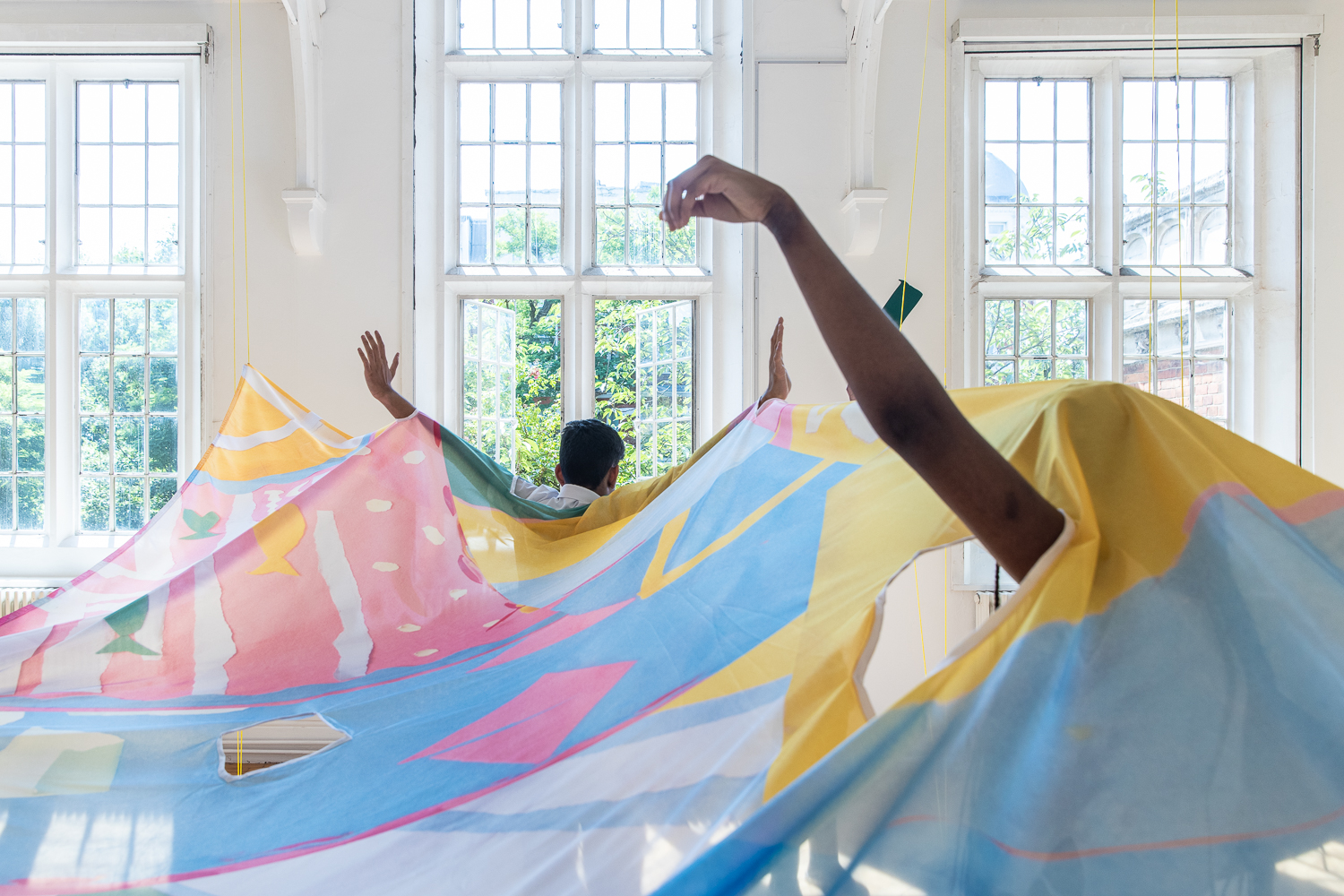 In front of white gallery windows, a fabric parachute is suspended from the ceiling. A child stands back towards it, arms up in the air. Another child's arm can be seen poking through a seamed hole.