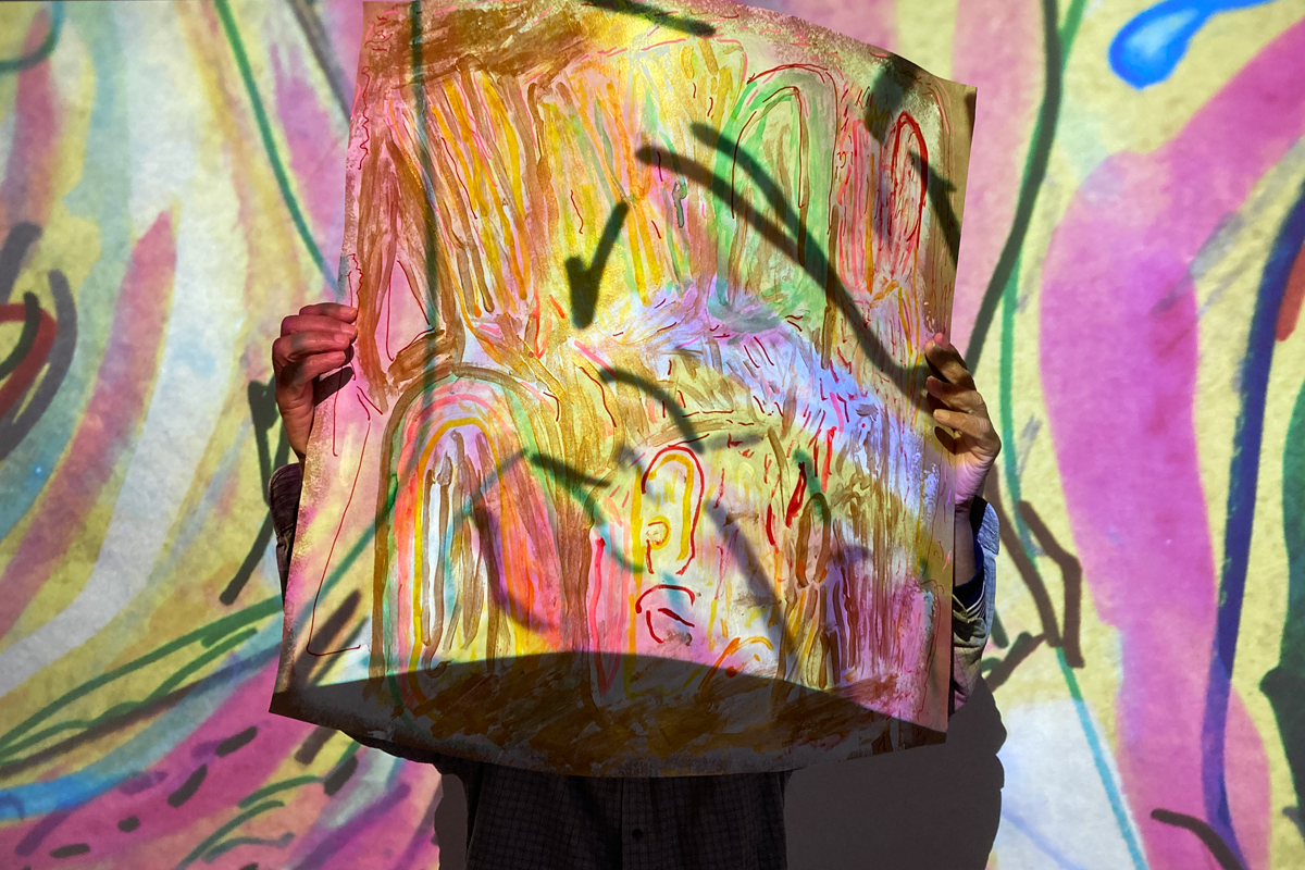 An artist holds up a drawing in front of their face. The wall is covered in a blown up version of his work and a projection is directed towards it.