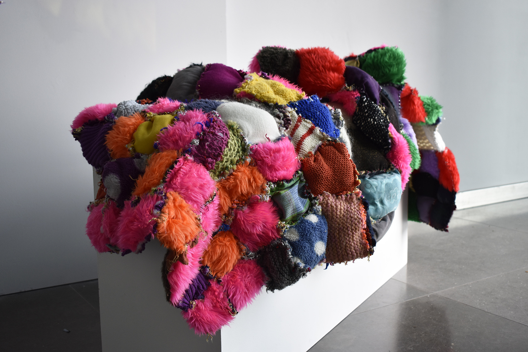Small squares of colourful fabric sewn together to create a large artwork that is draped over a white plinth.