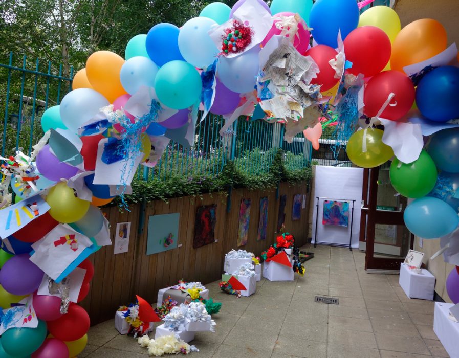 An outside space with concrete slabs and blue railings. It has lots of colourful balloons and white boxes placed around it.