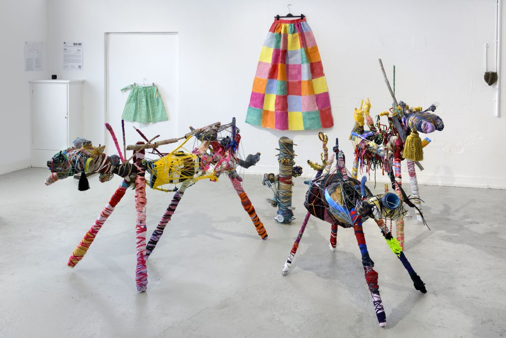Andrew's artwork stands in a gallery space. 3 of his sculptures are on the floor, which are all creatures made of different fabrics wound together with string. Behind them a colourful patchwork curtain hangs on a wall.