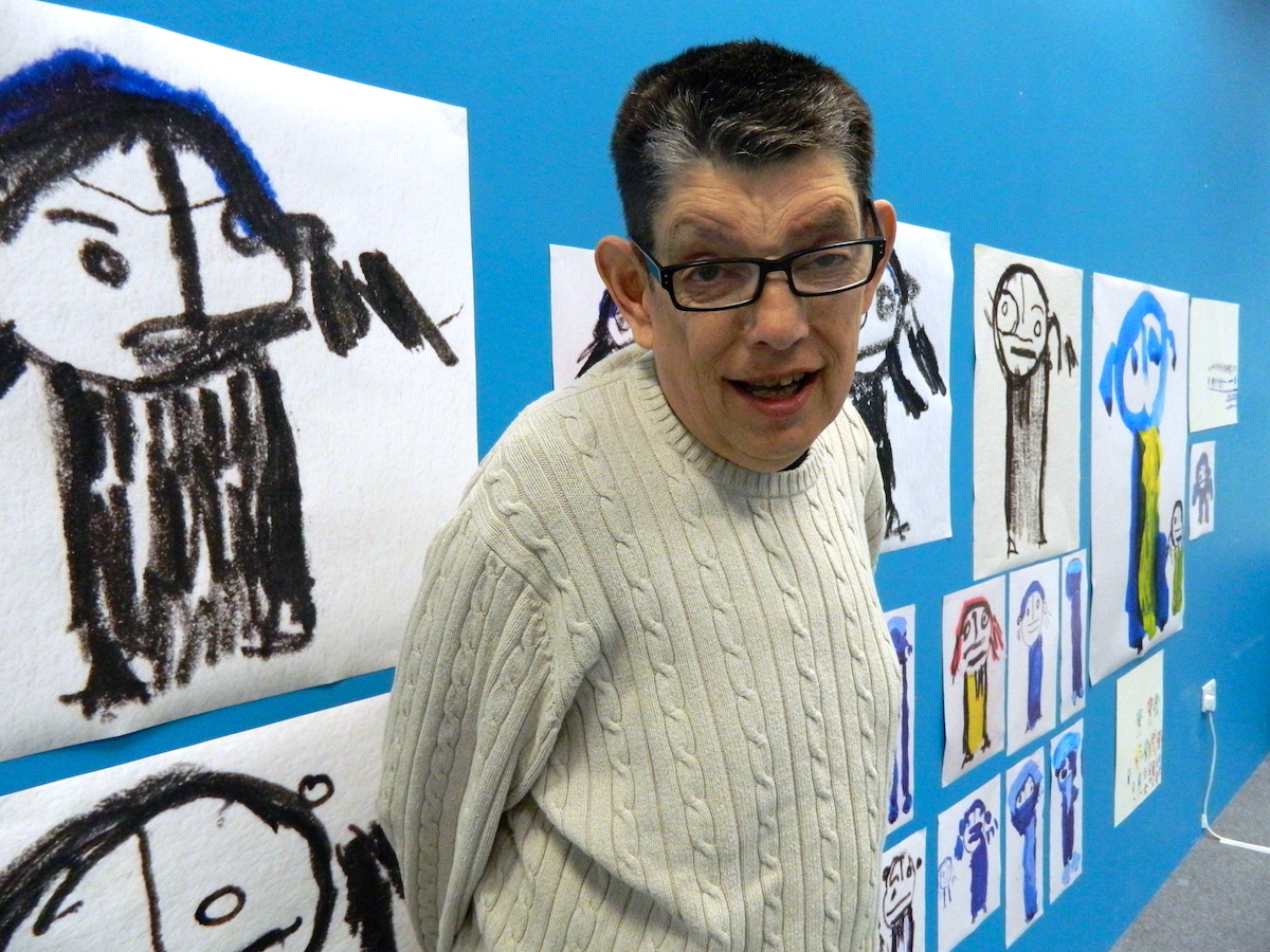 An image of Roland young, smiling at the camera. Behind him are his drawings on a blue wall. The drawings are charcoal drawings of figures on white paper.