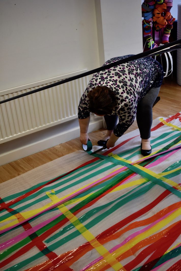 A woman bends down to stick colourful tape to the floor. There is a lot of it already stuck down in strips next to each other.