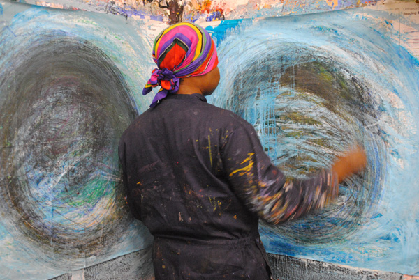 Nnena stands with her back to the camera, working on her drawings. There are two large pieces of paper in front of her with a blue, white and black scrawling circle on both.