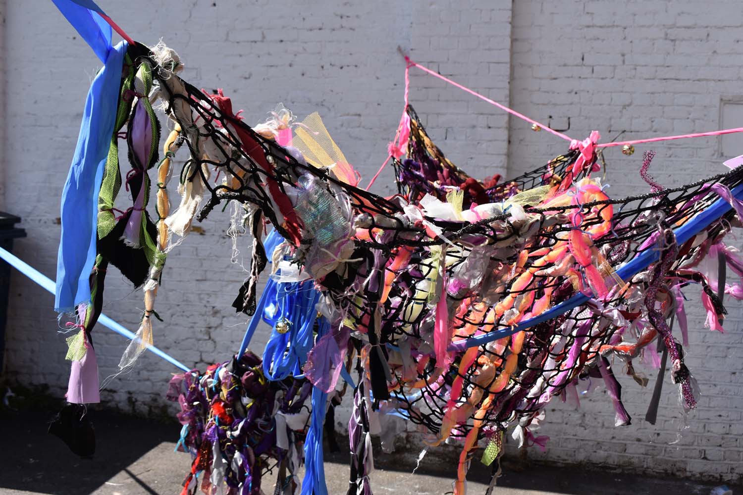 A suspended black net with different materials woven into it, hanging in a courtyard.