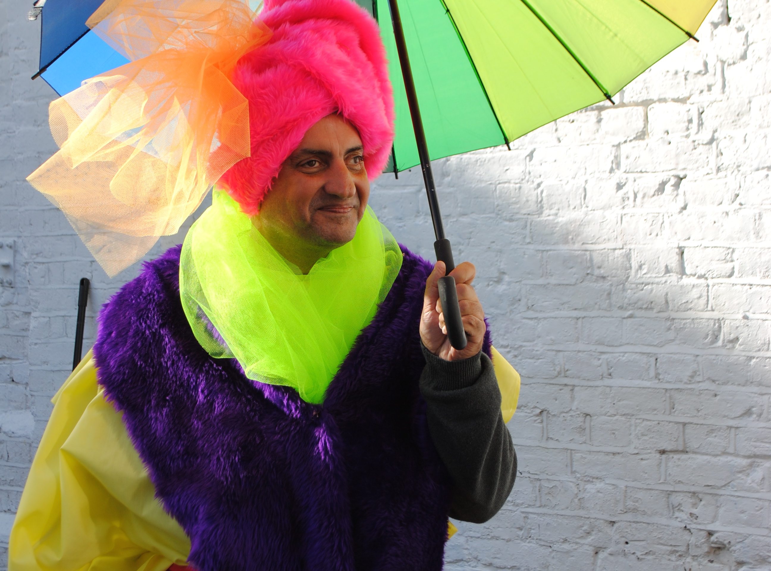 Artist Pardip Kapil, wearing a custom bright pink boa turban with blue and silver tinsel, and a yellow robe with a purple trim and neon green netted scarf, holding a rainbow umbrella.