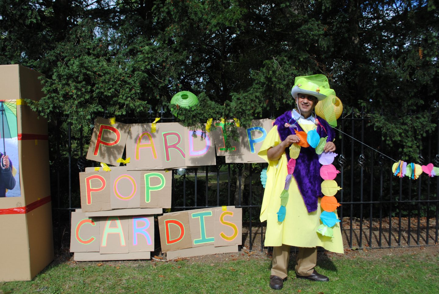 Pardip Kapil stood beside cardboard signs that read "Pardip Pop Cardis" attached to a black iron fence outdoors. He wears a DIY costume with a yellow robe and cowboy hat.
