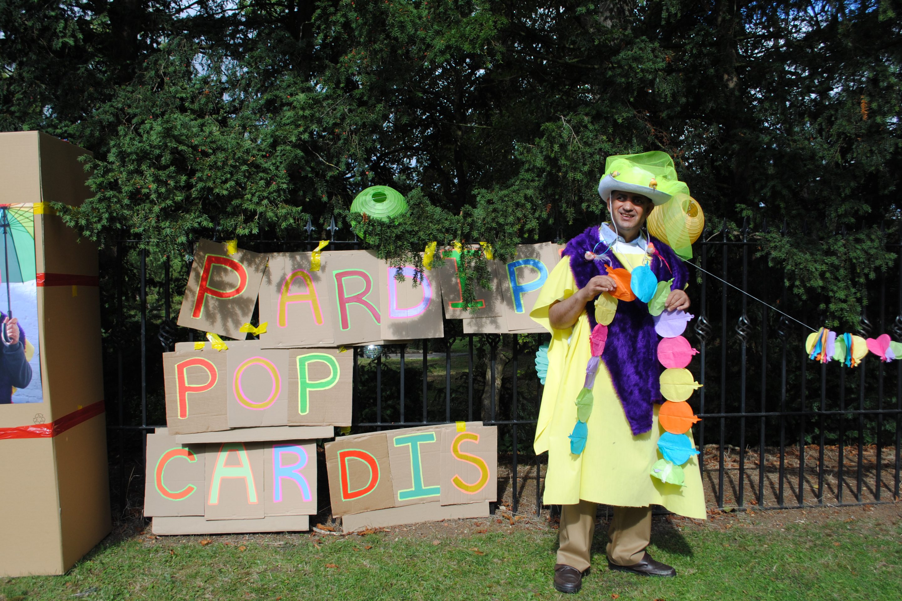 Pardip Kapil stood beside cardboard signs that read "Pardip Pop Cardis" attached to a black iron fence outdoors. He wears a DIY costume with a yellow robe and cowboy hat.