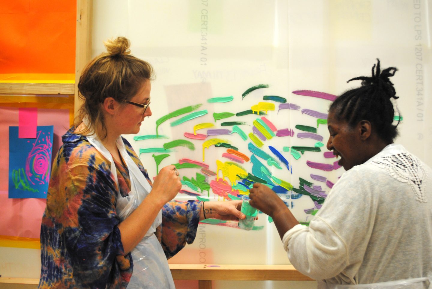 Emily Rees- Haynes with an artist. Emily is holding a pot with paint in, and the artist is dipping her brush in and painting the wall.