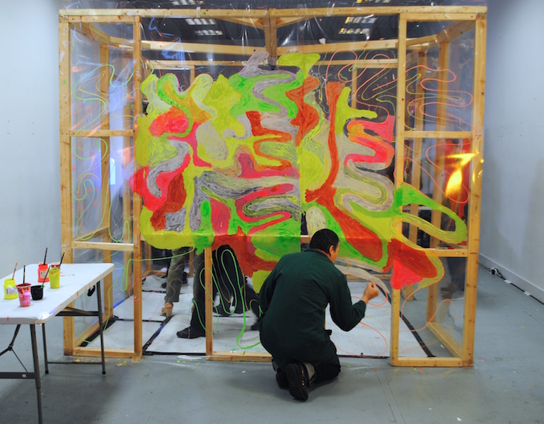 A person kneels down to draw on a large clear sheet that is stapled to a large wooden frame. Their drawing is abstract and colourful.