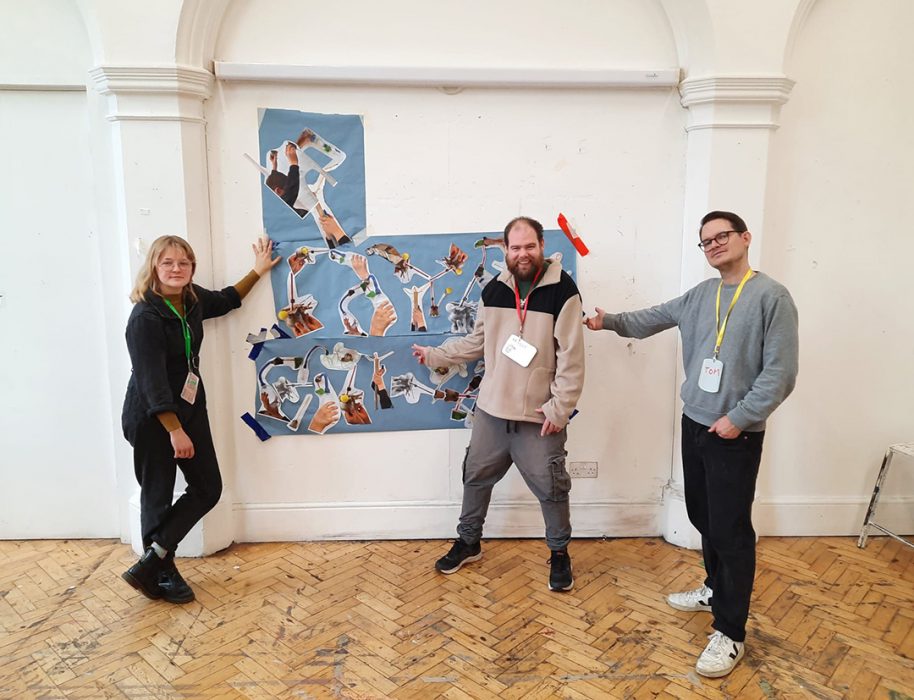 Declan Leslie stands in front of an artwork in a gallery space with two others. The work is large blue rolls of paper, with collaged photos stuck onto it of hands holding various objects.