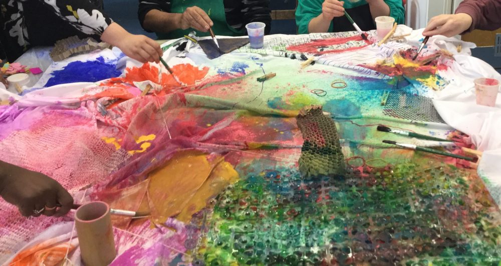 Several sheets of fabric are on a table, and people are painting on it.