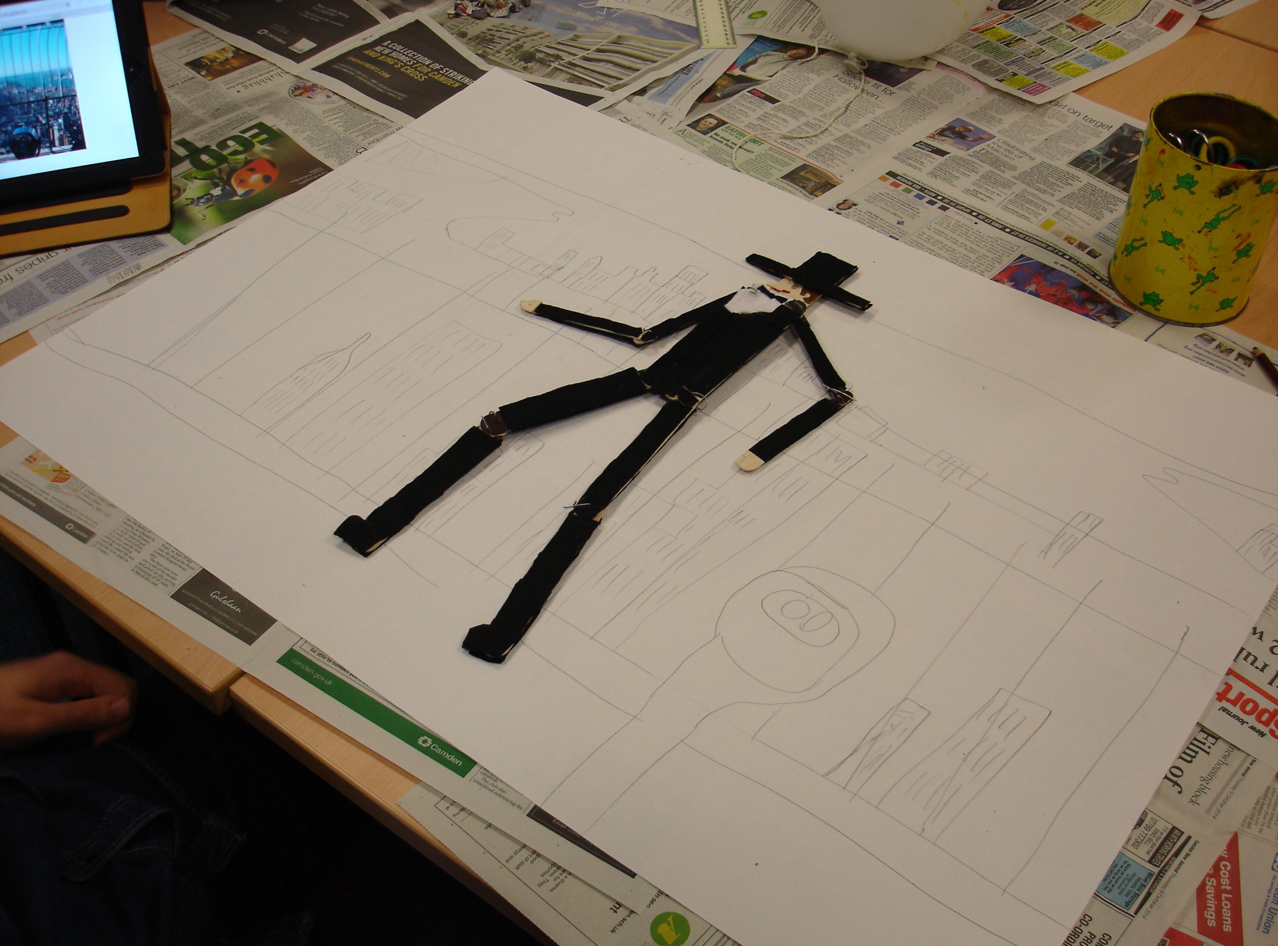 A figure of a man with a top hat made of lollipop sticks place upon a pencil drawing of a cityscape.
