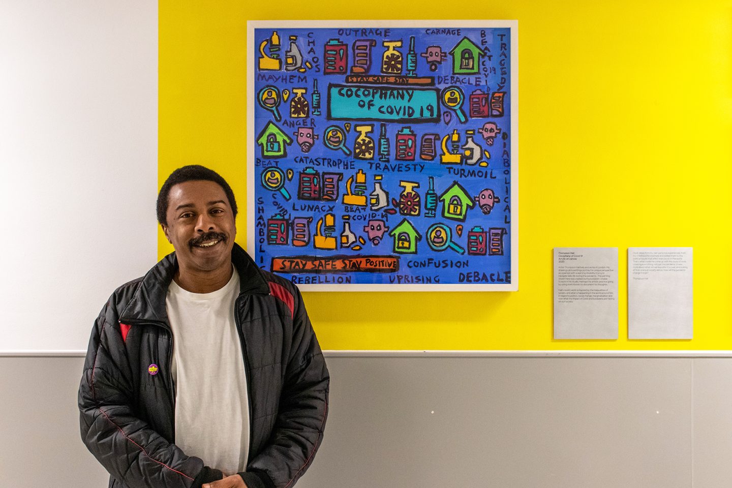 Thompson Hall stood in front of one of his artworks with a blue background and a white frame. Hung on a yellow gallery wall.