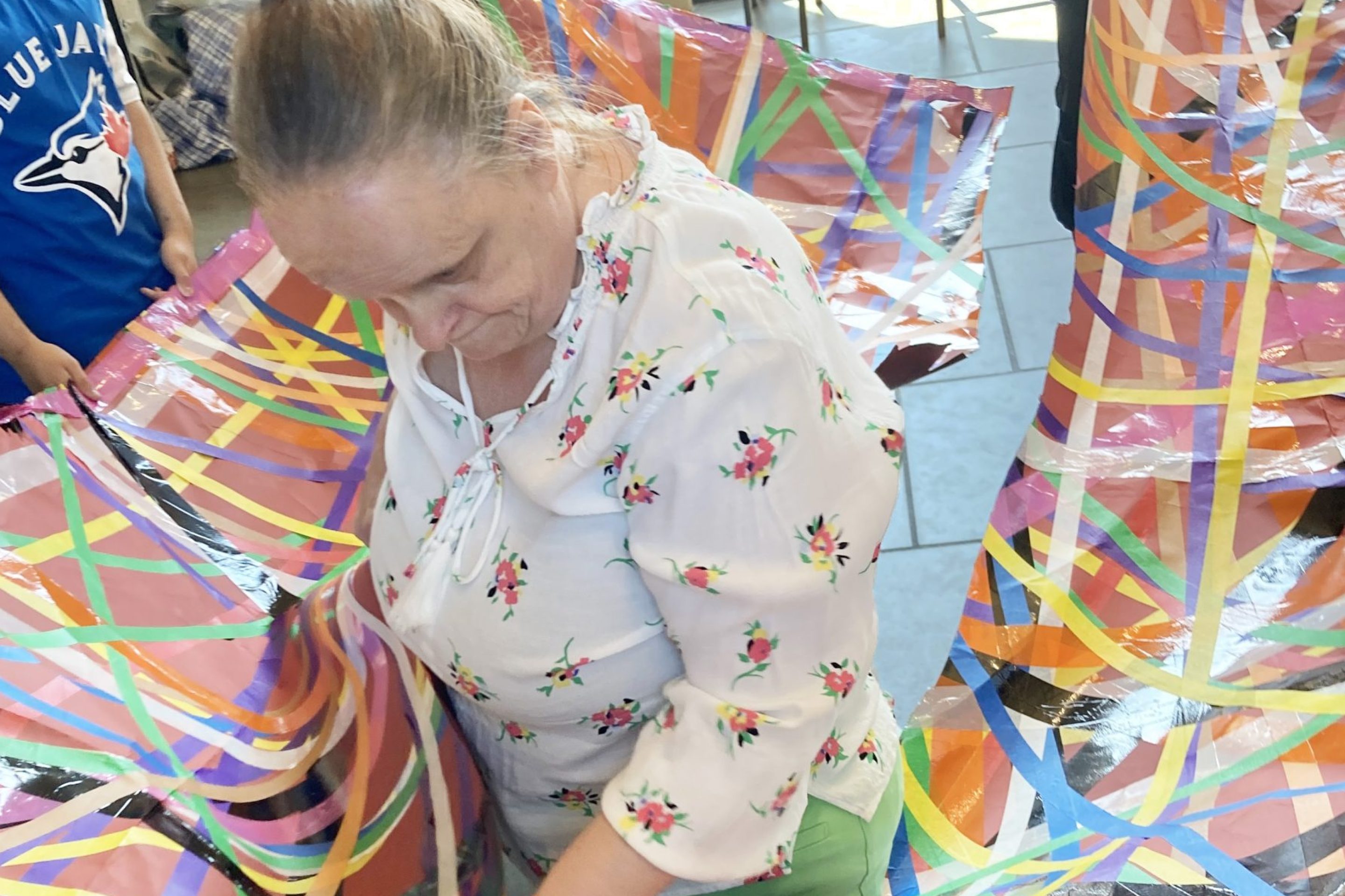 Linda Bell stands inside a large colourful sheet, with colourful tape all over it. Participants stand outside of the sheet, holding it at waist level.