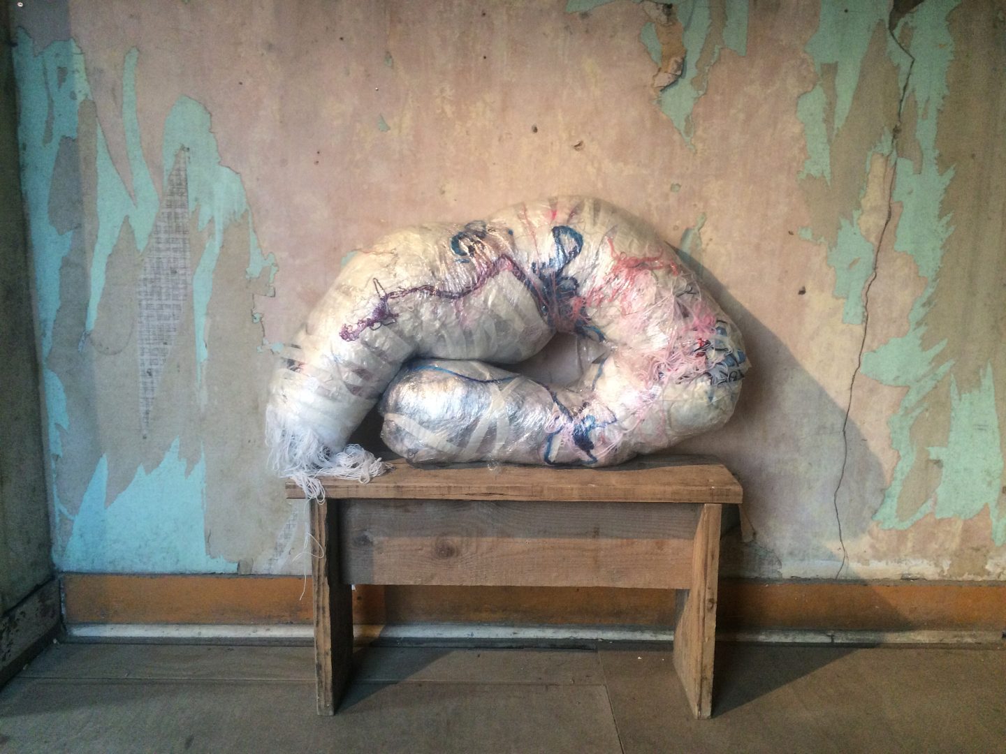 A multimedia sculpture, made of yarns and textiles wrapped in plastic, resting on a wooden bench.