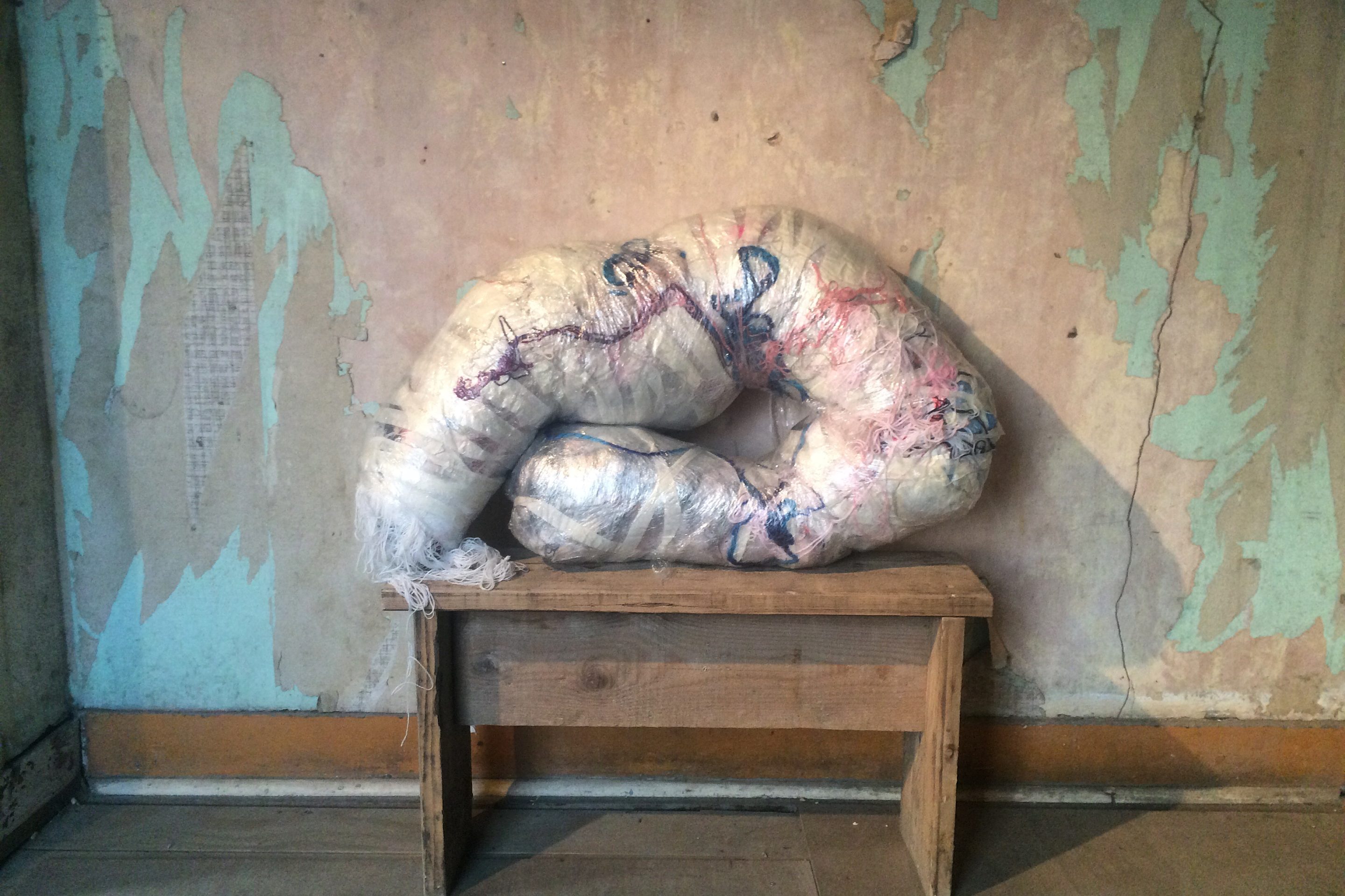 A multimedia sculpture, made of yarns and textiles wrapped in plastic, resting on a wooden bench.