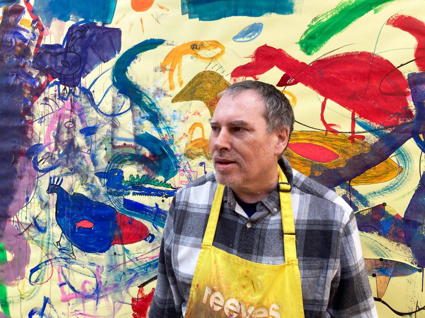 Robin Smith stands in front of a large artwork of his, wearing a yellow apron.